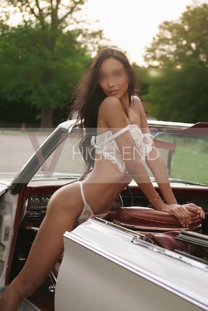 Sexy escort Thalia is leaning on the vintage car seat in a white lingerie