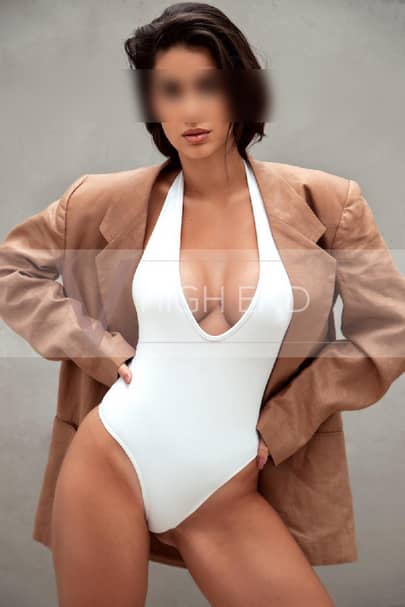 High-class escort Nina is holding her hips, dressed in a white bodysuit and jacket around her shoulders.
