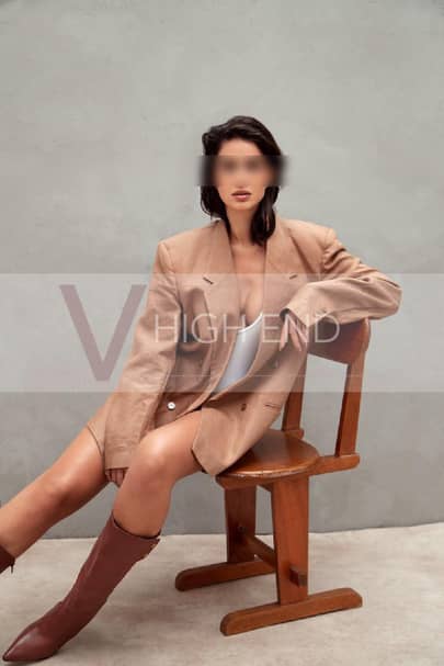 Sophisticated escort Nina dressed in a beige jacket and boots, sitting on a chair