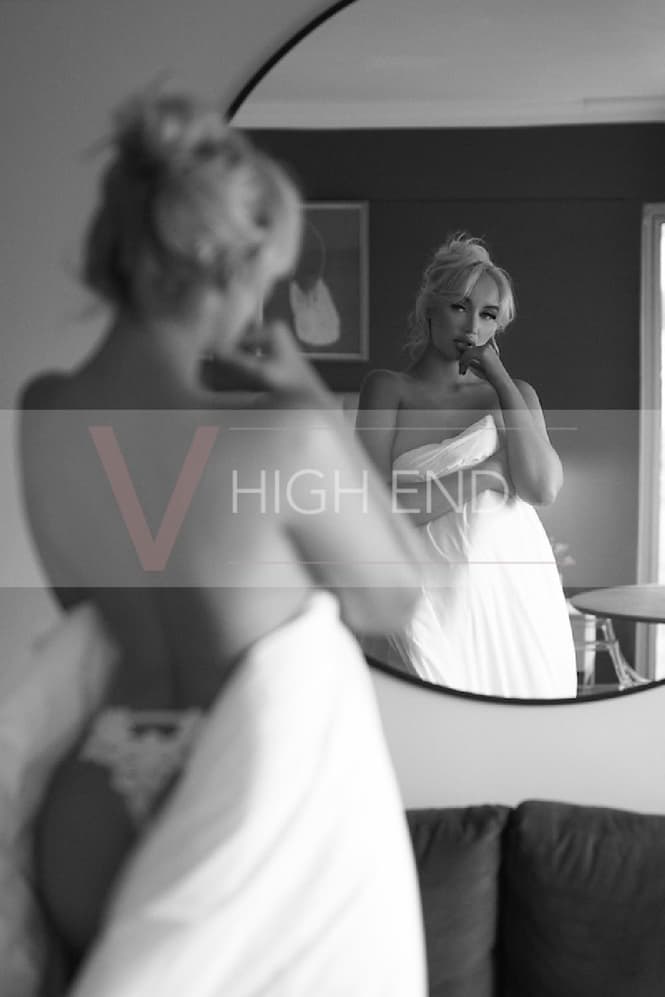 White and black photo of blonde escort Athena hiding her breast behind duvet