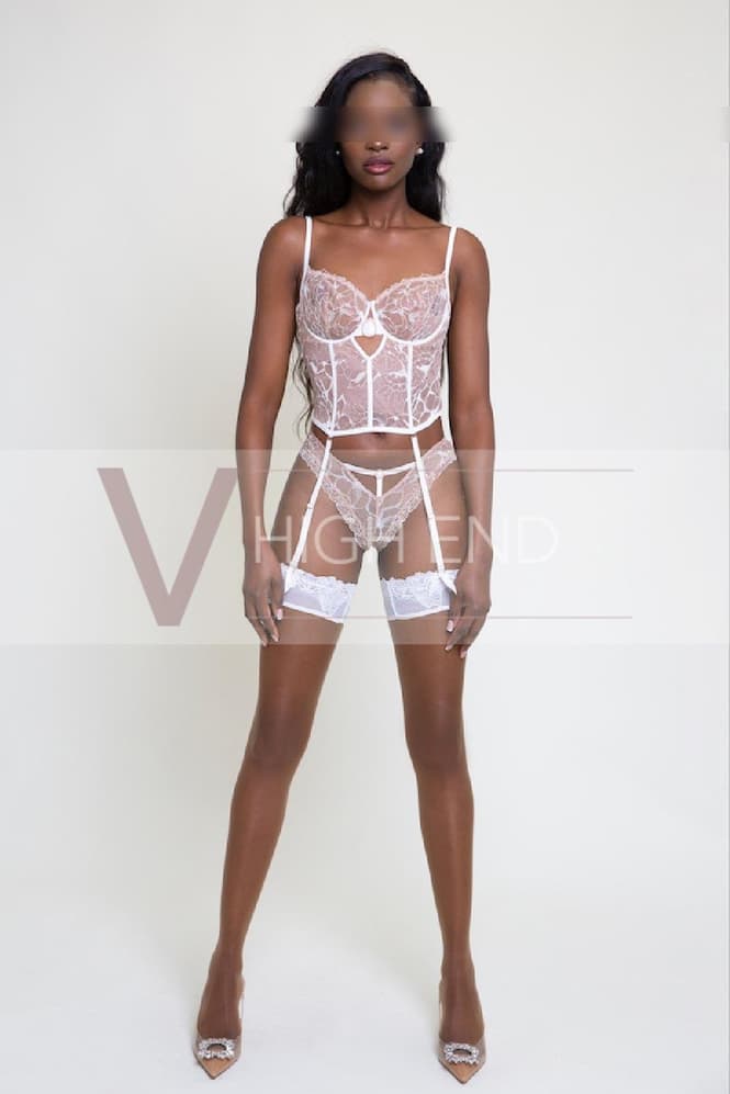 Premium escort Melanie posing on plain background wearing a white corset, matching panties, hold ups, suspenders belt and high heels shoes