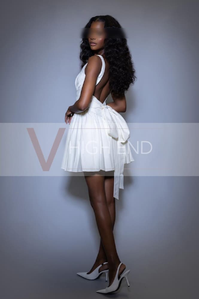 Sensual brunette companion Melanie weating white dress with a bow on her bum and white high heels shoes