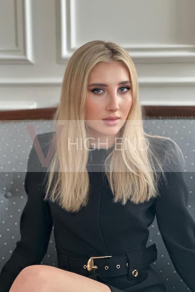 The blue-eyed model, Madelina, is sitting down in a black short outfit, giving a glimpse of her bare legs