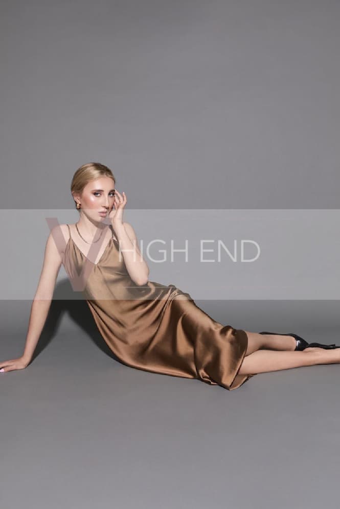 Tall escort Madelina, posing with a confident and natural posture, is wearing a gold dress, necklace, and earrings.