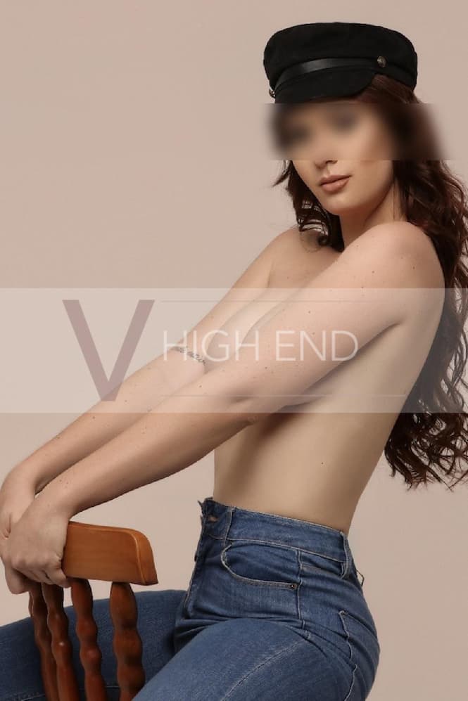 Topless escort Celina, dressed in jeans and a hat, is riding a chair.