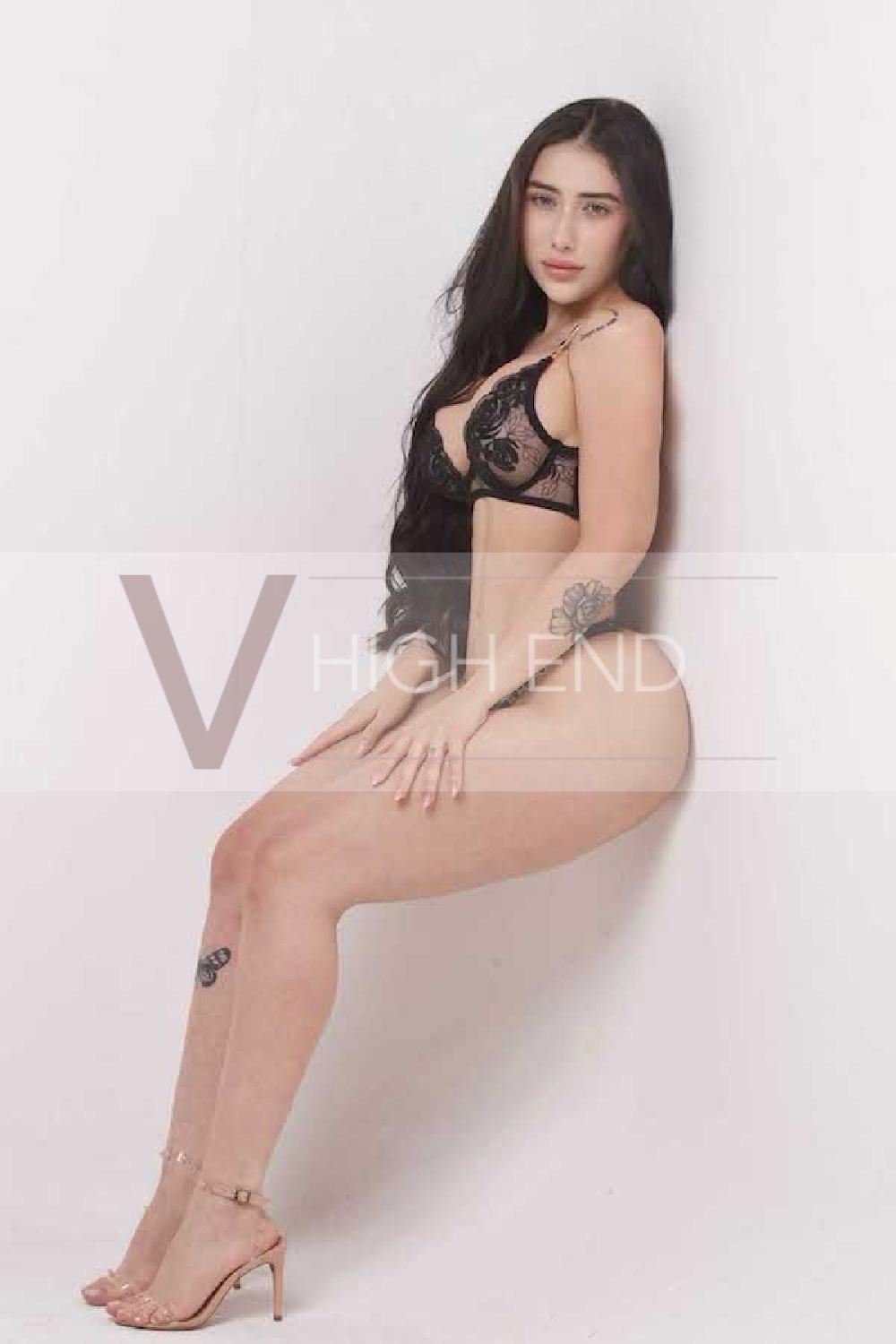 Vip escort Paulina is leaning on a white wall holding legs and wearing black underwear with beige high heels sandals 