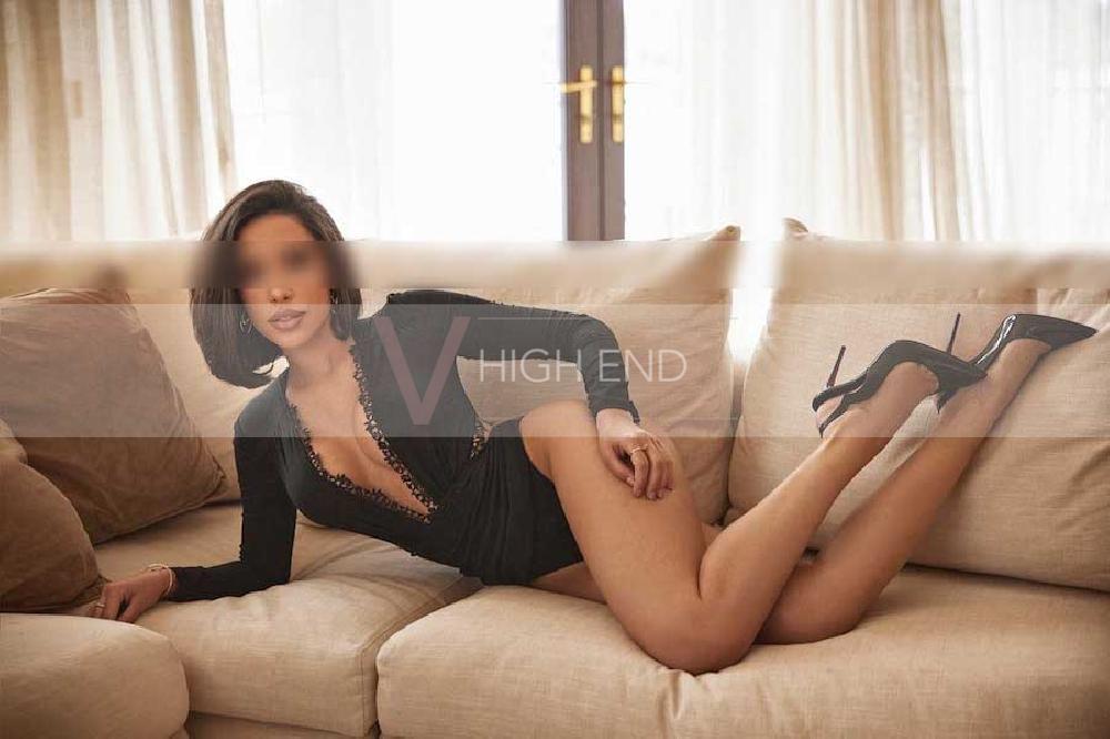 Brunette escort Iza is posing on the beige sofa wearing tiny black attire with her legs up